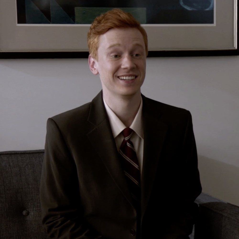 Kevin in a suit and tie, smiling on a sofa
